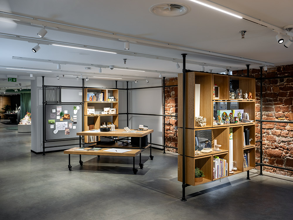 Modernes Interieur mit industriellen Elementen, offenen Regalen mit Büchern und Dekorationsartikeln, einem Holzschreibtisch mit Dokumenten, freiliegenden Backsteinwänden und Schienenbeleuchtung an der Decke.