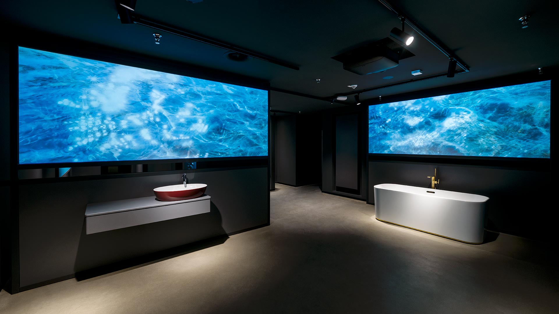 In einem modernen Zimmer mit dunklen Wänden sind ein rotes Waschbecken und eine weiße Badewanne zu sehen, beide unter großen beleuchteten Bildschirmen, die lebhafte Unterwasserszenen mit blauem Wasser zeigen.