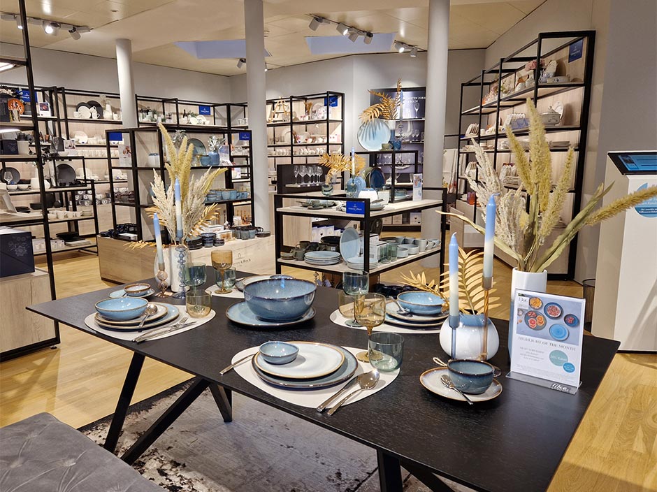 In einem modernen Ausstellungsraum wird ein Esstisch von Villeroy & Boch mit Dekor gezeigt, im Hintergrund stehen Regale mit Geschirr.
