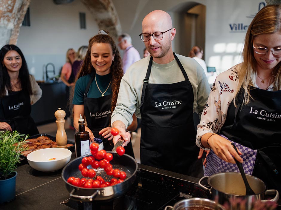Eine Gruppe von Menschen in Schürzen kocht gemeinsam in einer Küche. Ein Mann wirft Kirschtomaten in eine Pfanne, während andere lächeln und in Töpfen rühren und sich an der Tätigkeit erfreuen. Die Umgebung wirkt modern und gut beleuchtet.