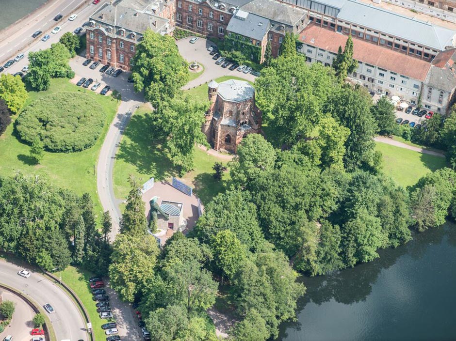 Luftaufnahme eines Parks mit dichten grünen Bäumen, einer gewundenen Straße, geparkten Autos, einem runden Gebäude in der Mitte (Alter Turm, Mettlach) und historischen Backsteinbauten in der Nähe eines Flusses.