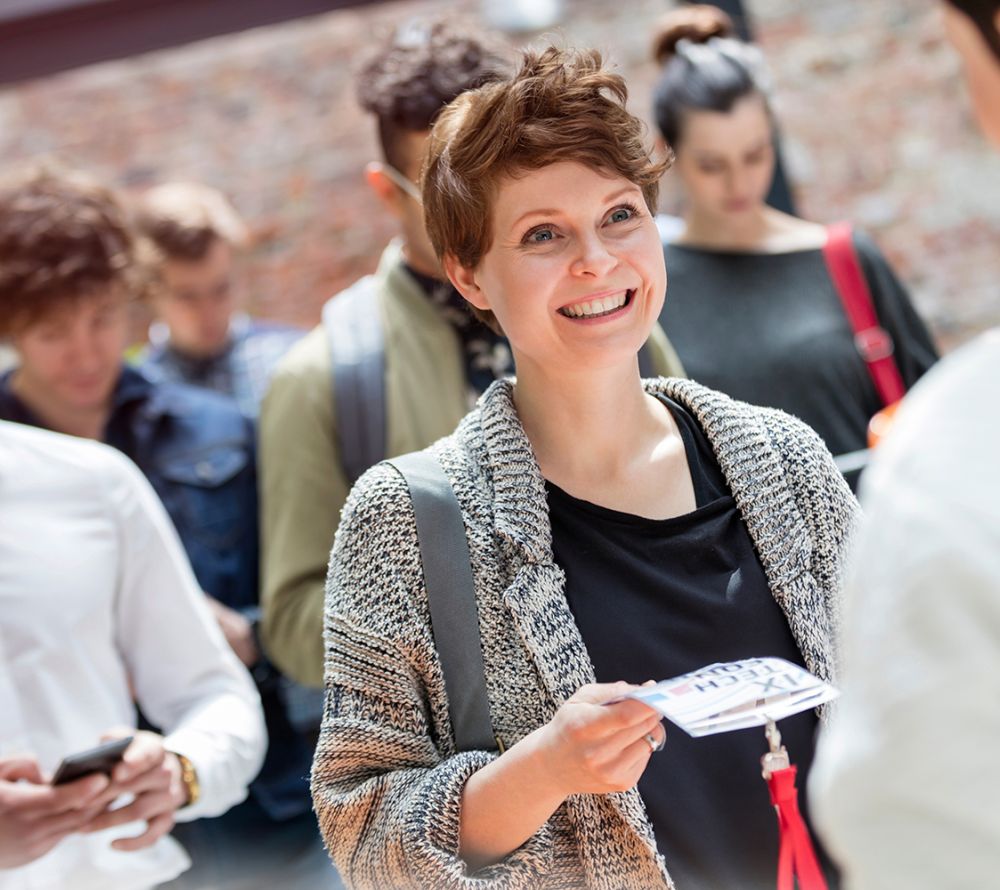 Eine lächelnde Frau mit einer Fahrkarte oder einem Pass steht draußen in einer Schlange, umgeben von anderen Menschen. Sie trägt eine graue Strickjacke und hat eine Tasche über der Schulter. Der Hintergrund ist leicht unscharf.