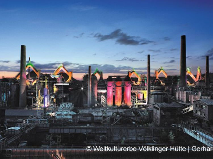 Ein Industriekomplex mit hohen Schornsteinen und bunt beleuchteten Strukturen, fotografiert in der Abenddämmerung mit einem dramatischen Himmel im Hintergrund. Der Text am unteren Rand verweist auf das Weltkulturerbe Völklinger Hütte und Gerhard.