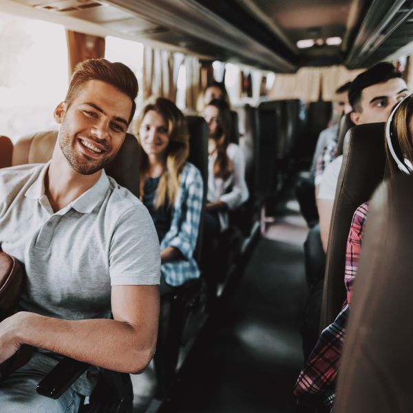 Eine Gruppe junger Erwachsener sitzt in einem Bus, lächelt und unterhält sich. Der Fokus liegt auf einem fröhlichen Mann im Vordergrund, der ein helles Poloshirt trägt. Im Hintergrund sind mehrere Personen zu sehen, die alle entspannt und glücklich wirken.