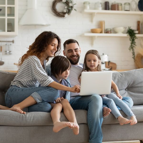 Eine lächelnde vierköpfige Familie sitzt auf einer Couch in einem gemütlichen Wohnzimmer und schaut gemeinsam auf einen Laptop. Die Eltern und zwei Kinder scheinen glücklich und beschäftigt zu sein und die gemeinsame Zeit zu genießen.