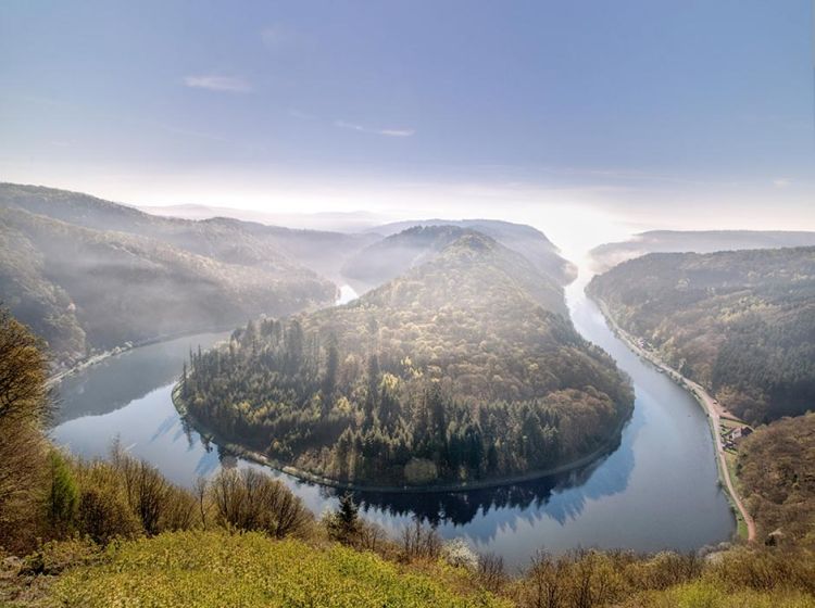 Ein breiter Fluss schlängelt sich malerisch um einen dicht bewaldeten Hügel, umgeben von nebelverhangenen, baumbestandenen Hängen unter klarem Himmel, die eine ruhige Naturlandschaft schaffen.